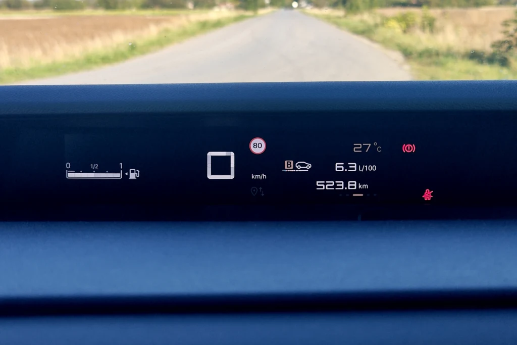 Wyświetlacz cyfrowych zegarów jest niewysoki i znajduje się pod samą przednią szybą Cyfrowy wyświetlacz samochodowy z informacjami o aktualnej prędkości, limicie dozwolonej prędkości, temperaturze zewnętrznej, wskaźniku paliwa, zużyciu paliwa oraz przewidywanym zasięgu, na tle widoku drogi i pól po obu stronach.