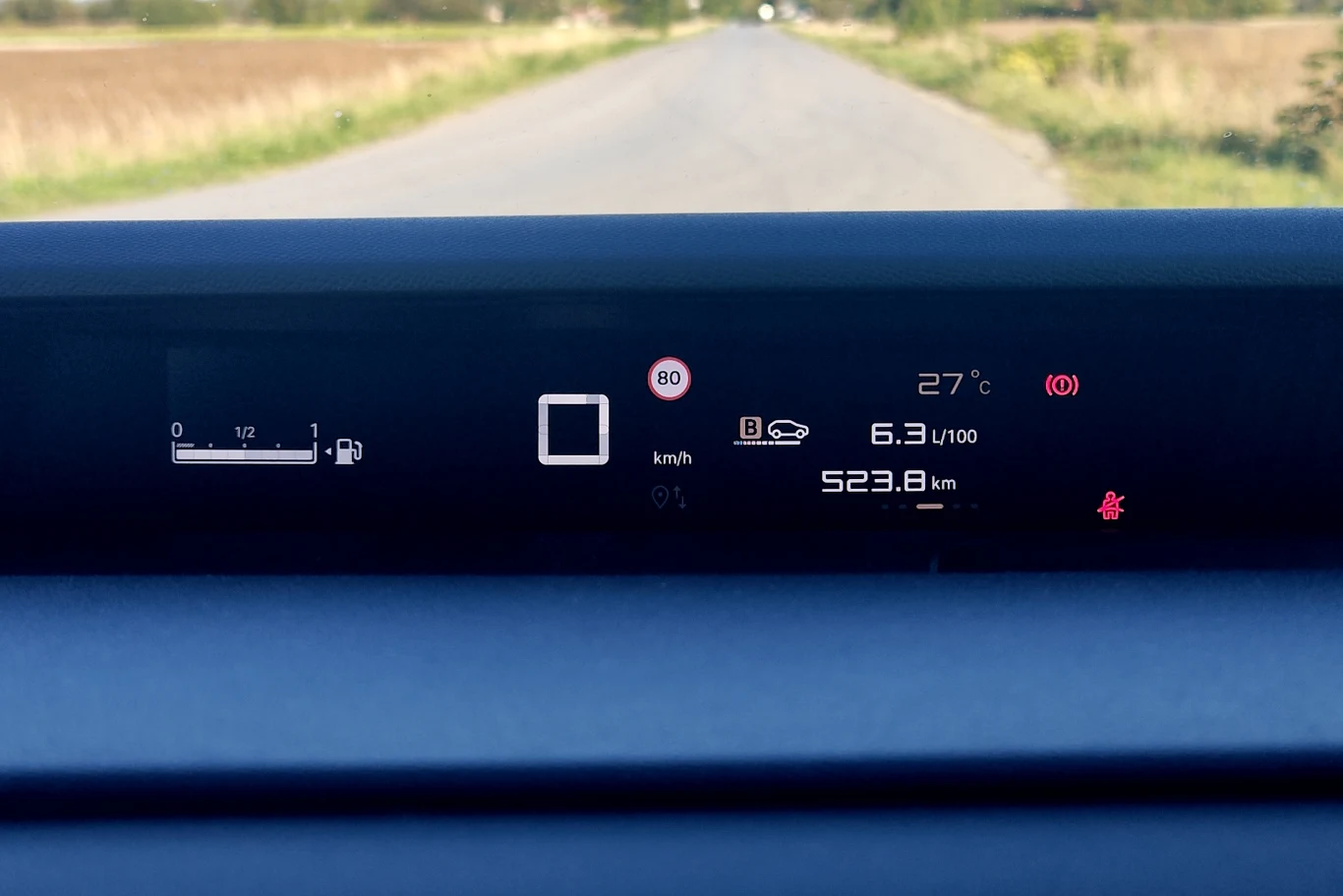 Cyfrowy wyświetlacz samochodowy z informacjami o aktualnej prędkości, limicie dozwolonej prędkości, temperaturze zewnętrznej, wskaźniku paliwa, zużyciu paliwa oraz przewidywanym zasięgu, na tle widoku drogi i pól po obu stronach.