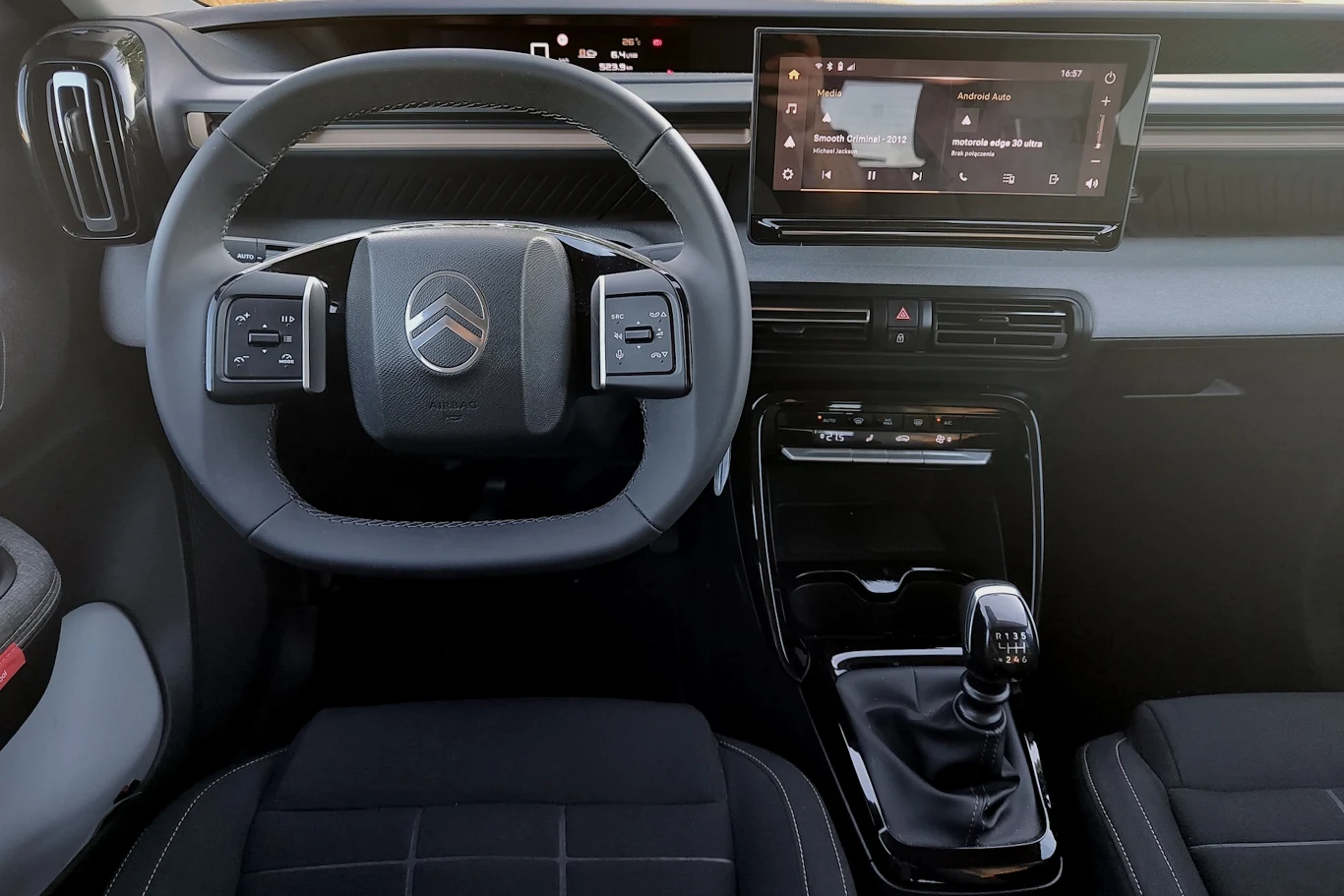 Kierownica samochodu Citroën, szeroka konsola z nowoczesnym wyświetlaczem dotykowym na środku, manualna skrzynia biegów oraz czarne, wygodne fotele w przedniej części kokpitu.