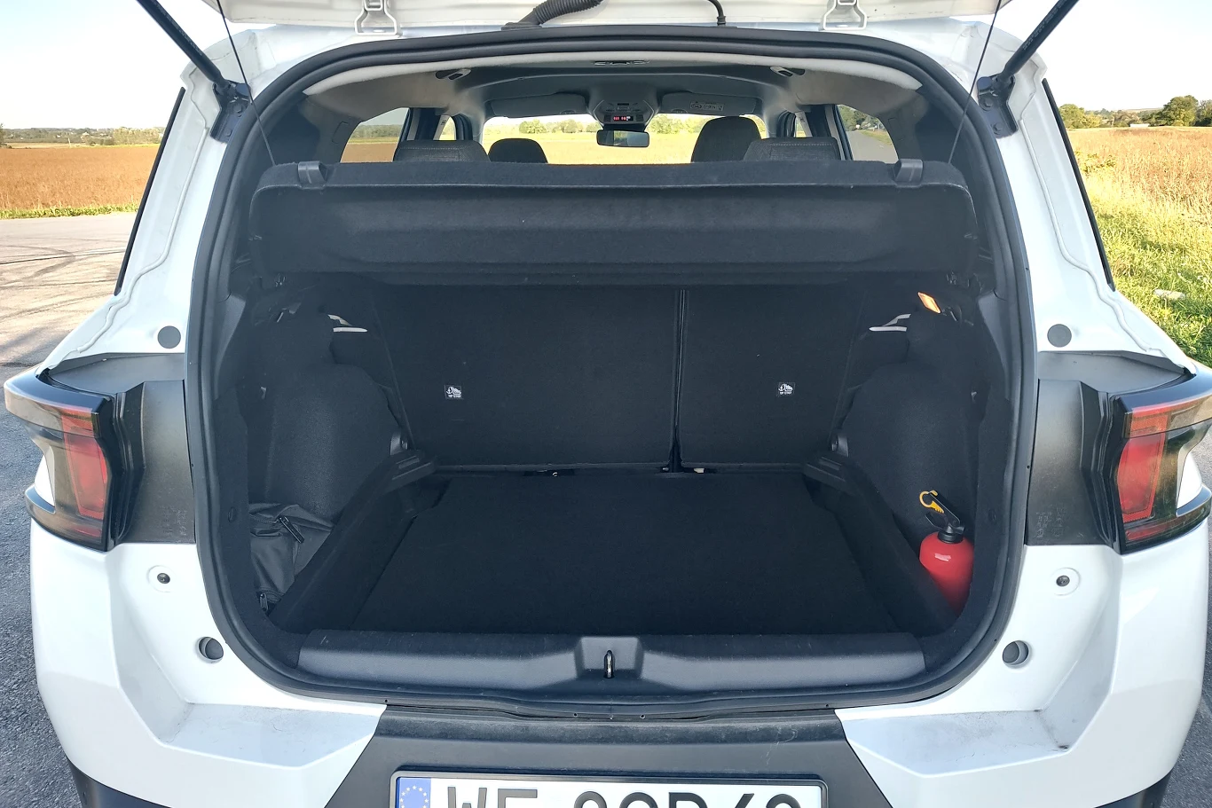 Otwarte wnętrze bagażnika białego samochodu osobowego z widoczną czarną wykładziną, składaną tylną kanapą oraz umieszczoną po prawej stronie butlą gaśnicy. W tle widok na pole pod jasnym niebem.