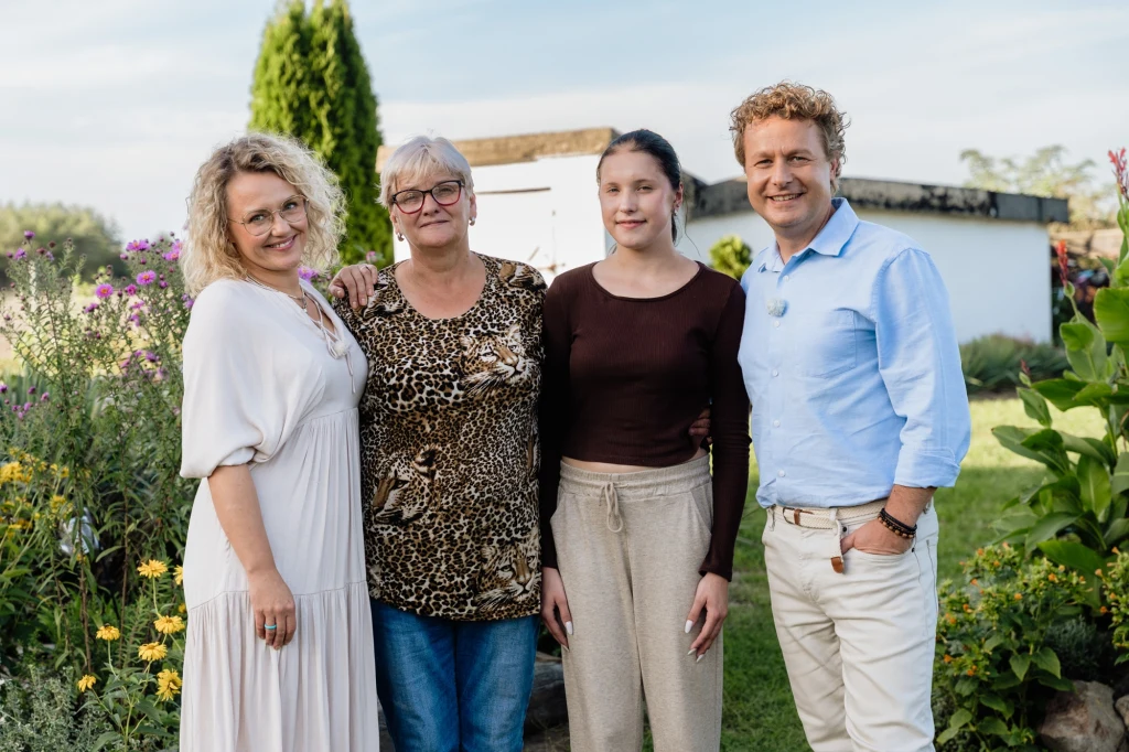 Cztery osoby pozujące razem w ogrodzie, otoczone wysokimi i kolorowymi kwiatami. W tle widoczny biały budynek oraz zielone drzewa. Osoby uśmiechnięte, stoją blisko siebie, tworząc rodzinny i przyjazny klimat.