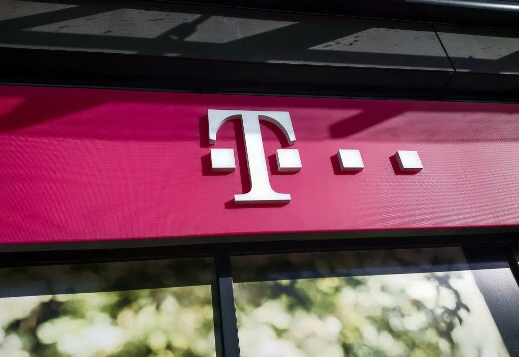 Logo T-Mobile na różowym tle, umieszczone na froncie budynku. Napisy są w kolorze białym, a w tle widoczne delikatne odbicia drzew i nieba na szybie.