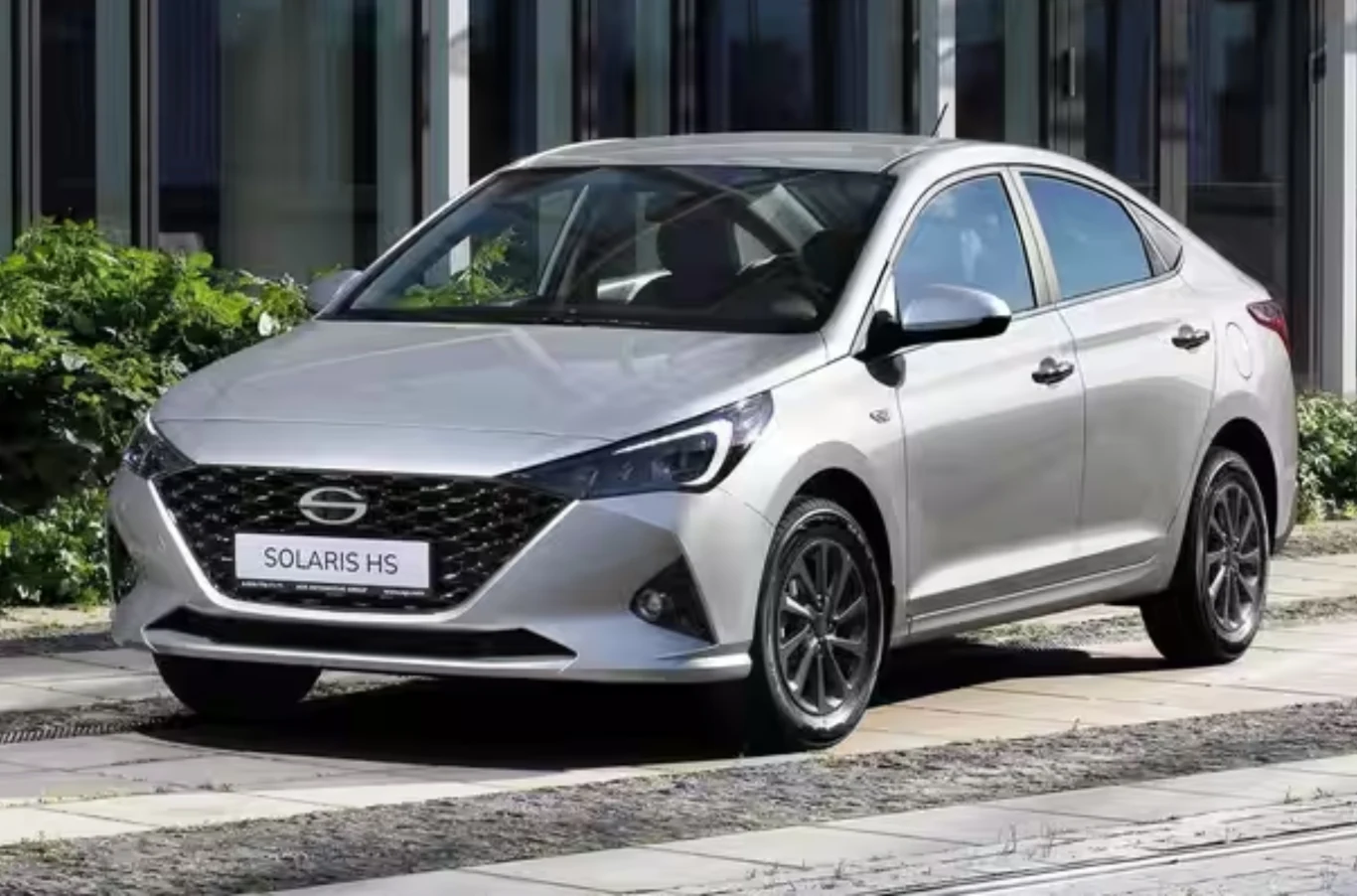 Srebrny sedan Solaris HS zaparkowany na tle nowoczesnego budynku, otoczony roślinnością i brukowanym chodnikiem, z widocznymi szczegółami przedniego grilla oraz felg.