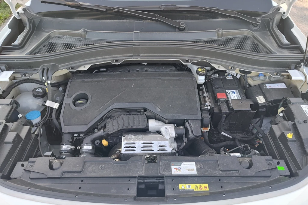 Silnik Citroena C3 Aircross ma 1,2 l pojemności i dzięki turbo generuje 100 KM. Elektryfikacji brak Komora silnika samochodu osobowego z widocznymi podzespołami takimi jak silnik, akumulator, wlewy płynów eksploatacyjnych oraz elementy elektryczne, przykryte częściowo plastikowymi osłonami.