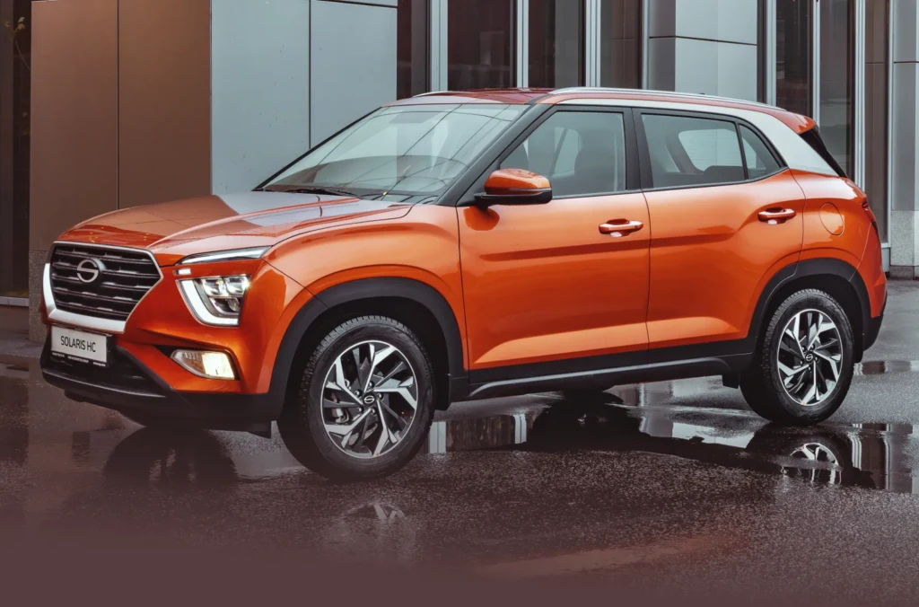 Pomarańczowy SUV zaparkowany przed nowoczesnym budynkiem o przeszklonych ścianach. Samochód stoi na mokrej nawierzchni i jest dobrze widoczny z profilu, ukazując nowoczesny design, aluminiowe felgi oraz charakterystyczne reflektory.