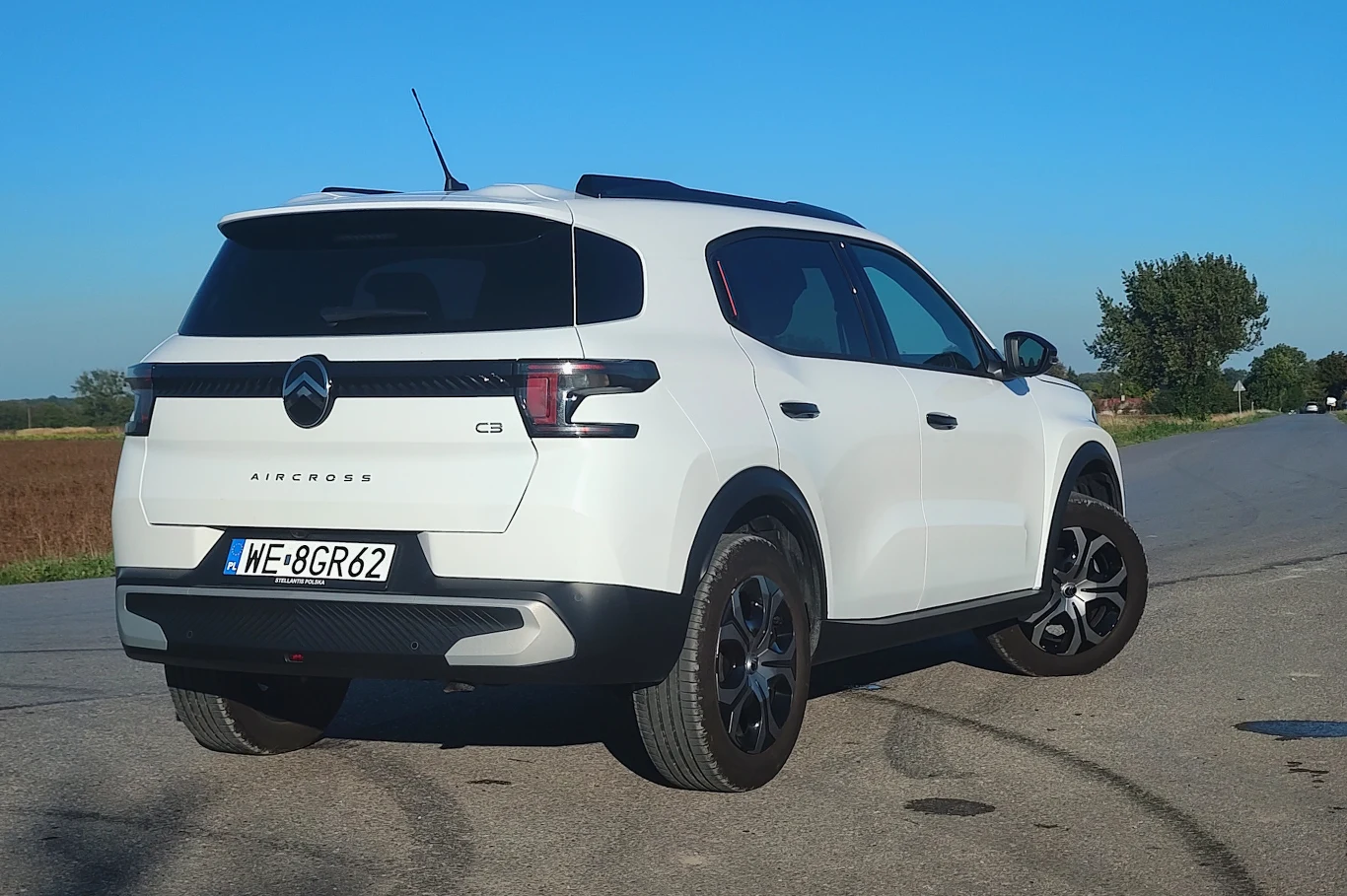 Biały samochód Citroën C5 Aircross stoi na wyludnionej asfaltowej drodze, otoczony polami i drzewami. Auto jest widoczne od tyłu, a na tablicy rejestracyjnej widnieje numer WE 8GR62. W tle błękitne niebo bez chmur.