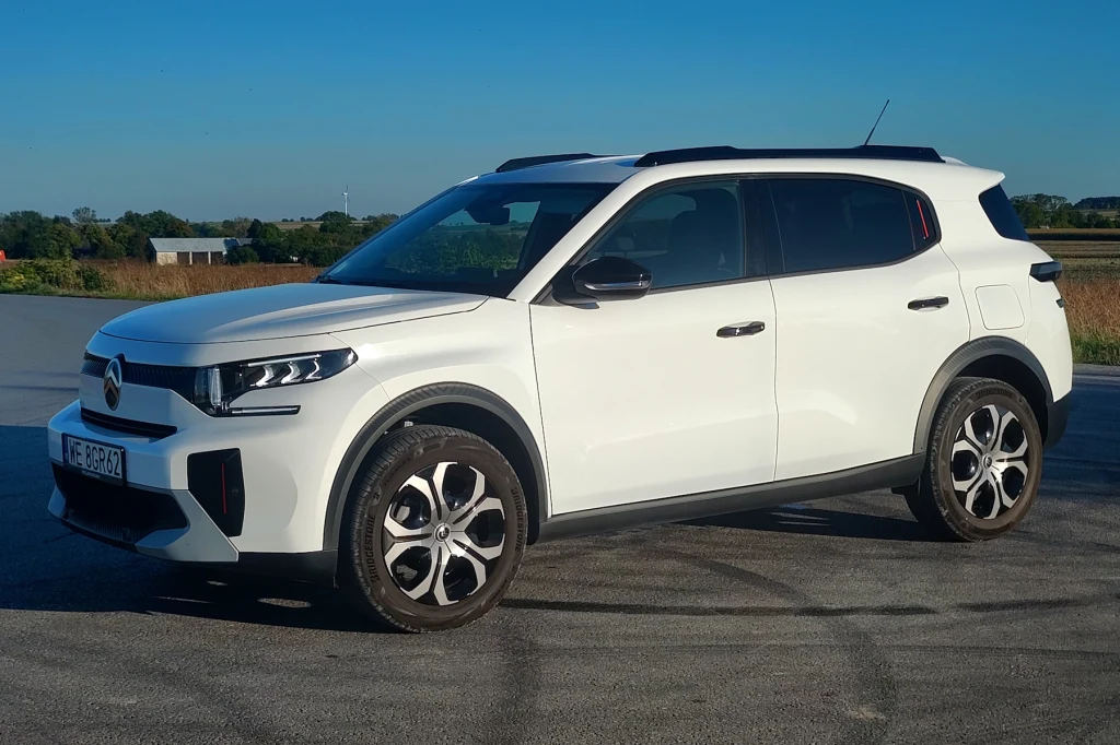 Citroen C3 Aircross jest dostępny również jako hybryda MHEV i auto elektryczne Biały samochód typu SUV stoi na asfaltowej drodze, na tle pola i niebieskiego nieba, z widocznymi nowoczesnymi reflektorami i charakterystycznymi felgami.