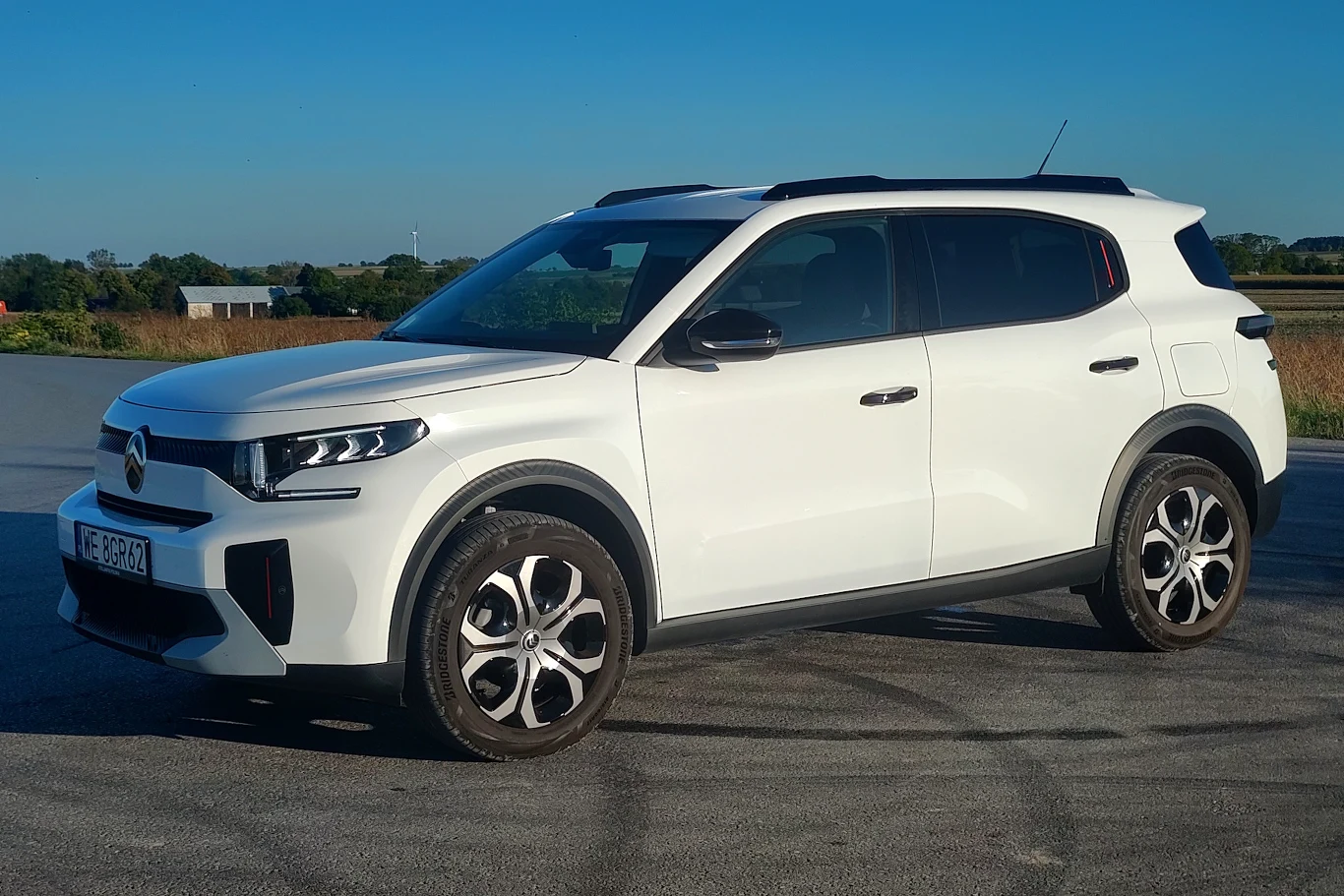 Biały samochód typu SUV stoi na asfaltowej drodze, na tle pola i niebieskiego nieba, z widocznymi nowoczesnymi reflektorami i charakterystycznymi felgami.