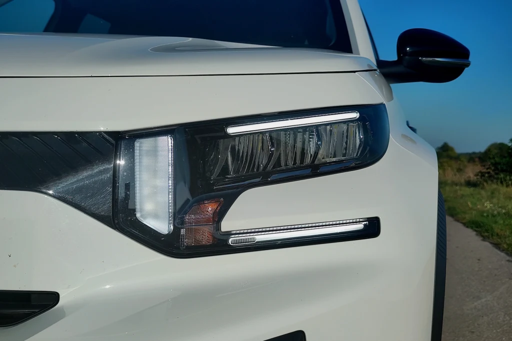 Citroen C3 Aircross Zbliżenie na przedni reflektor białego samochodu typu SUV z wyraźnym kształtem świateł i fragmentem maski, w tle niebieskie niebo oraz roślinność przy drodze.