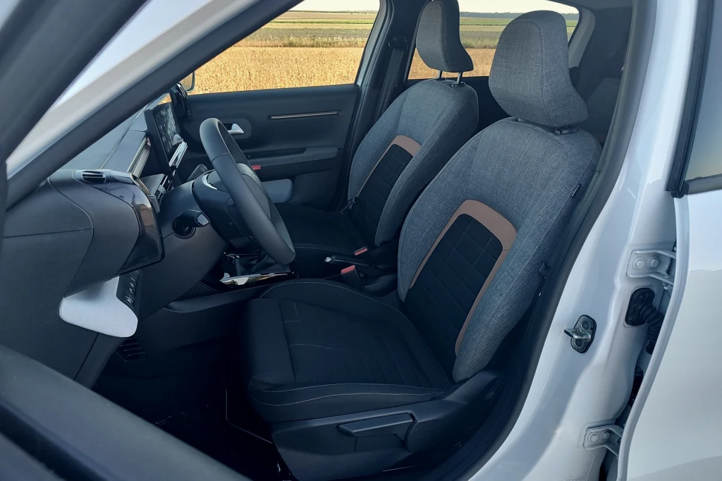Fotele są wygodne, mogą być podgrzewane, ale sterowanie jest manualne Wnętrze samochodu z widocznymi dwoma przednimi siedzeniami, deską rozdzielczą oraz fragmentem panelu sterowania. Fotele pokryte są materiałową tapicerką w odcieniach szarości i czerni, z brązowymi akcentami. Przez otwarte drzwi auta widać krajobraz z p...