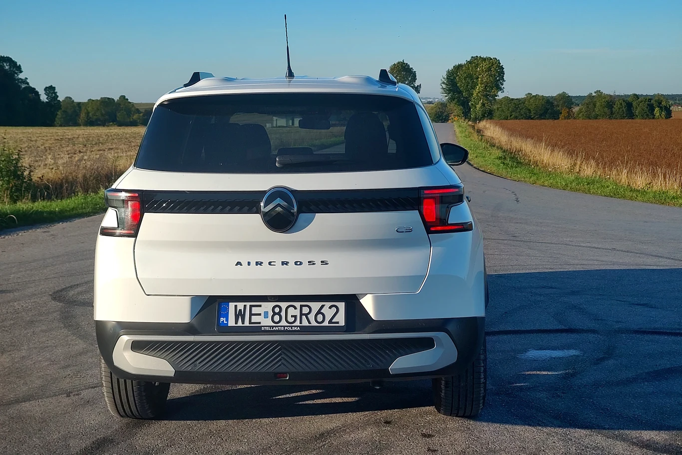 Biały samochód Citroën Aircross widziany od tyłu, stoi zaparkowany na wiejskiej drodze z płaskimi polami po obu stronach. Jasne światło podkreśla detale tylnej części auta. Otoczenie tworzą pola uprawne oraz drzewa przy horyzoncie.