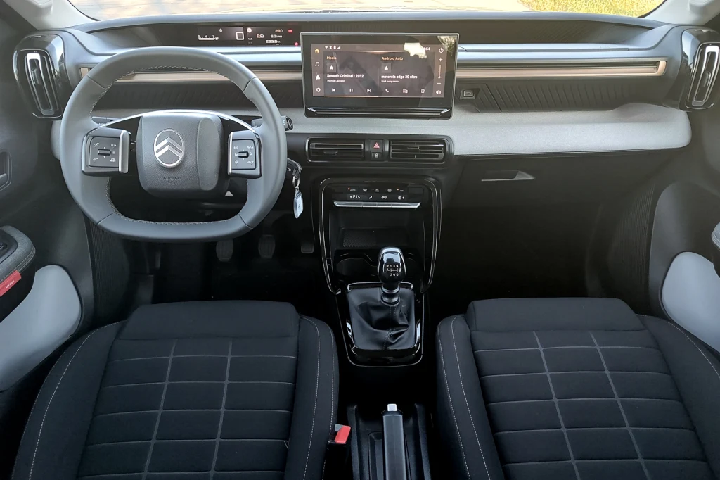 Citroen C3 Aircross Kokpit nowoczesnego samochodu marki Citroën z widocznym kierownicą, wyświetlaczem multimedialnym na środku deski rozdzielczej oraz dwoma przednimi fotelami pokrytymi czarną tapicerką. W centralnej części znajduje się dźwignia zmiany biegów oraz panel s...