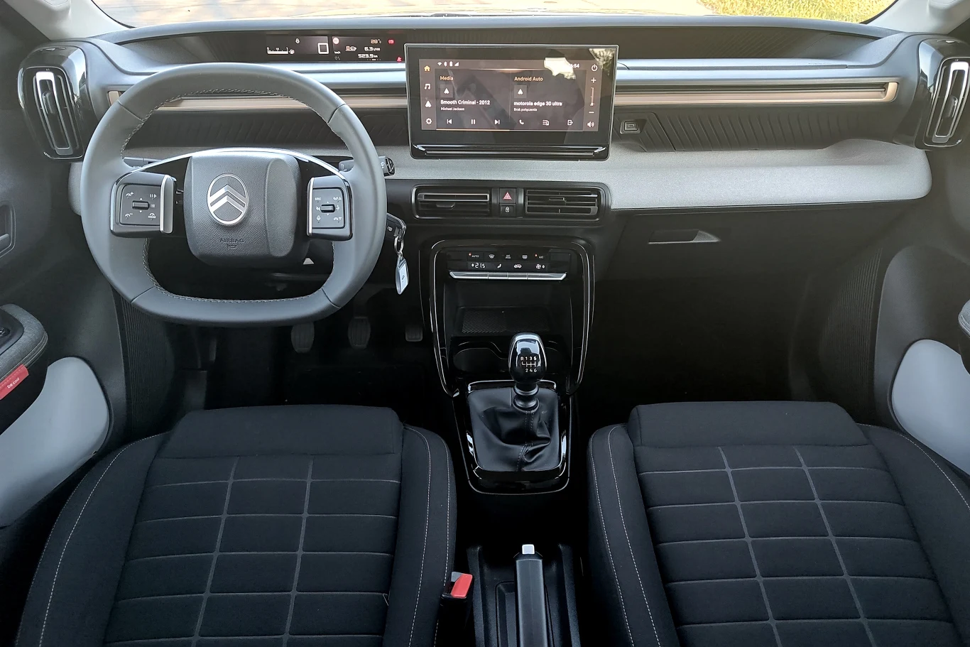 Kokpit nowoczesnego samochodu marki Citroën z widocznym kierownicą, wyświetlaczem multimedialnym na środku deski rozdzielczej oraz dwoma przednimi fotelami pokrytymi czarną tapicerką. W centralnej części znajduje się dźwignia zmiany biegów oraz panel s...