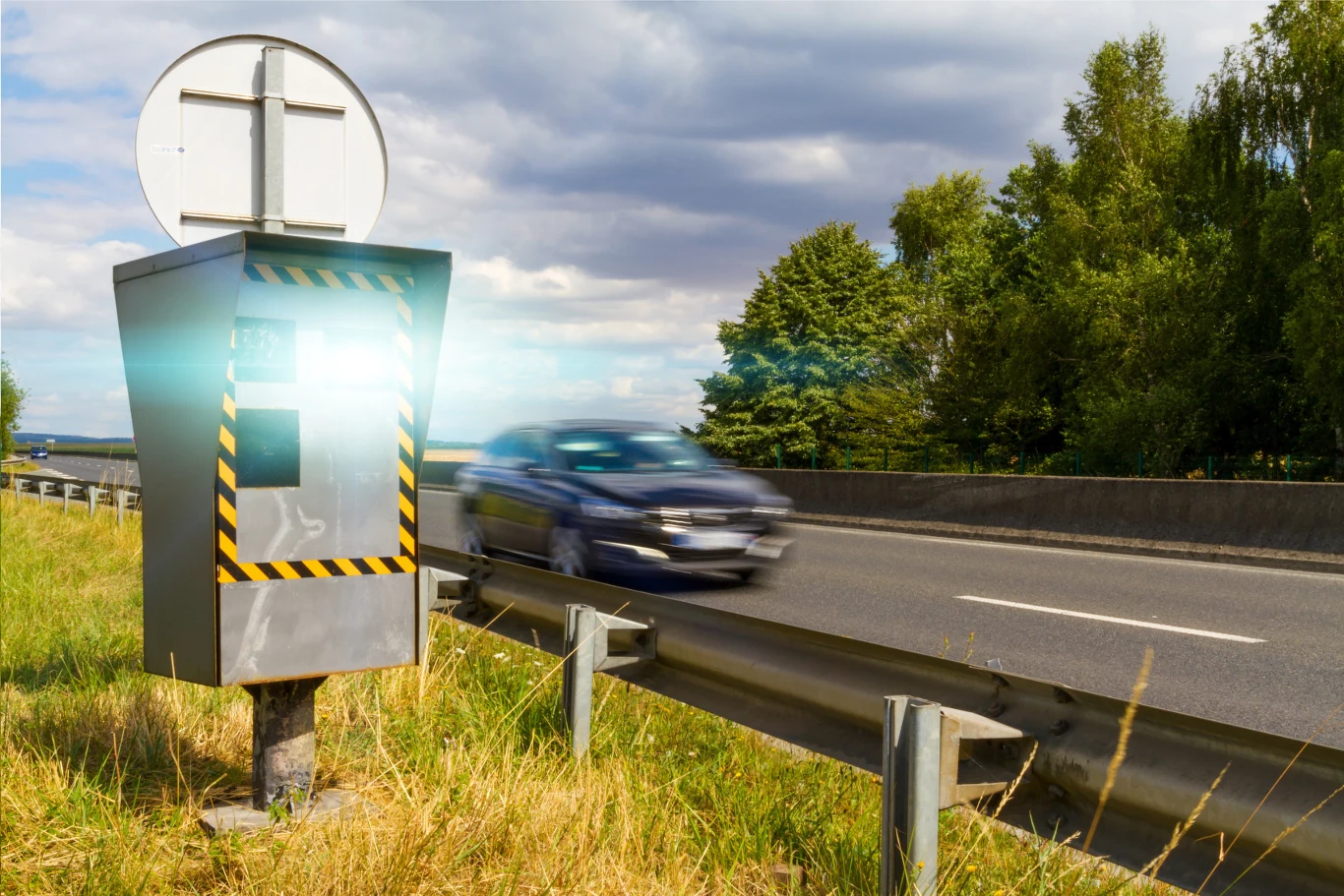 Kierowcy masowo dostają mandat z fotoradaru za jazdę z prędkością zgodną z obowiązującym limitem na drodze RD903. Fotoradar wykonujący zdjęcie przejeżdżającemu z dużą prędkością samochodowi na drodze osłoniętej barierkami, w otoczeniu zieleni oraz pochmurnego nieba.