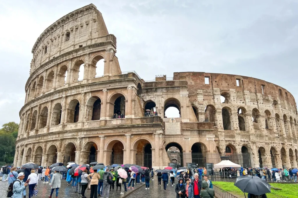 Koloseum w Rzymie otoczone przez liczną grupę turystów, z których wielu trzyma parasole. Rzymski amfiteatr dominuje w kadrze, a na pierwszym planie widać ludzi spacerujących wokół zabytku.