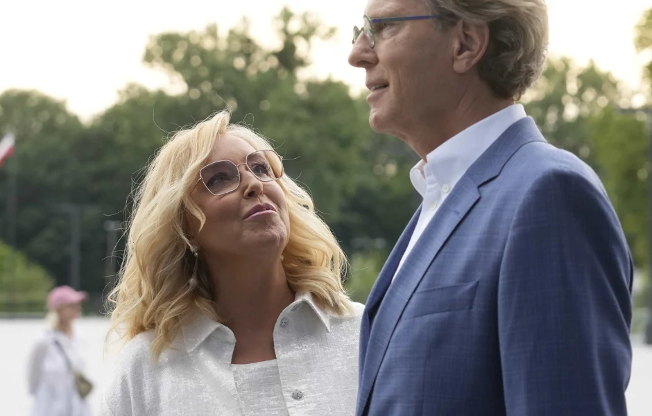 Dwie elegancko ubrane osoby stoją na świeżym powietrzu, rozmawiając ze sobą. Kobieta w jasnej marynarce patrzy z uśmiechem na mężczyznę w niebieskiej marynarce. W tle drzewa i zamazana sylwetka innej osoby.