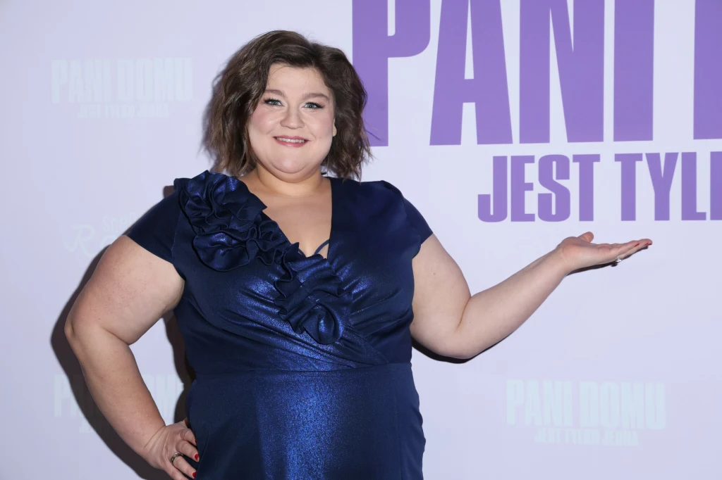 Uśmiechnięta kobieta ubrana w granatową sukienkę z kwiatową aplikacją stoi na tle plakatu i wykonuje gest wskazujący ręką na napis po prawej stronie.