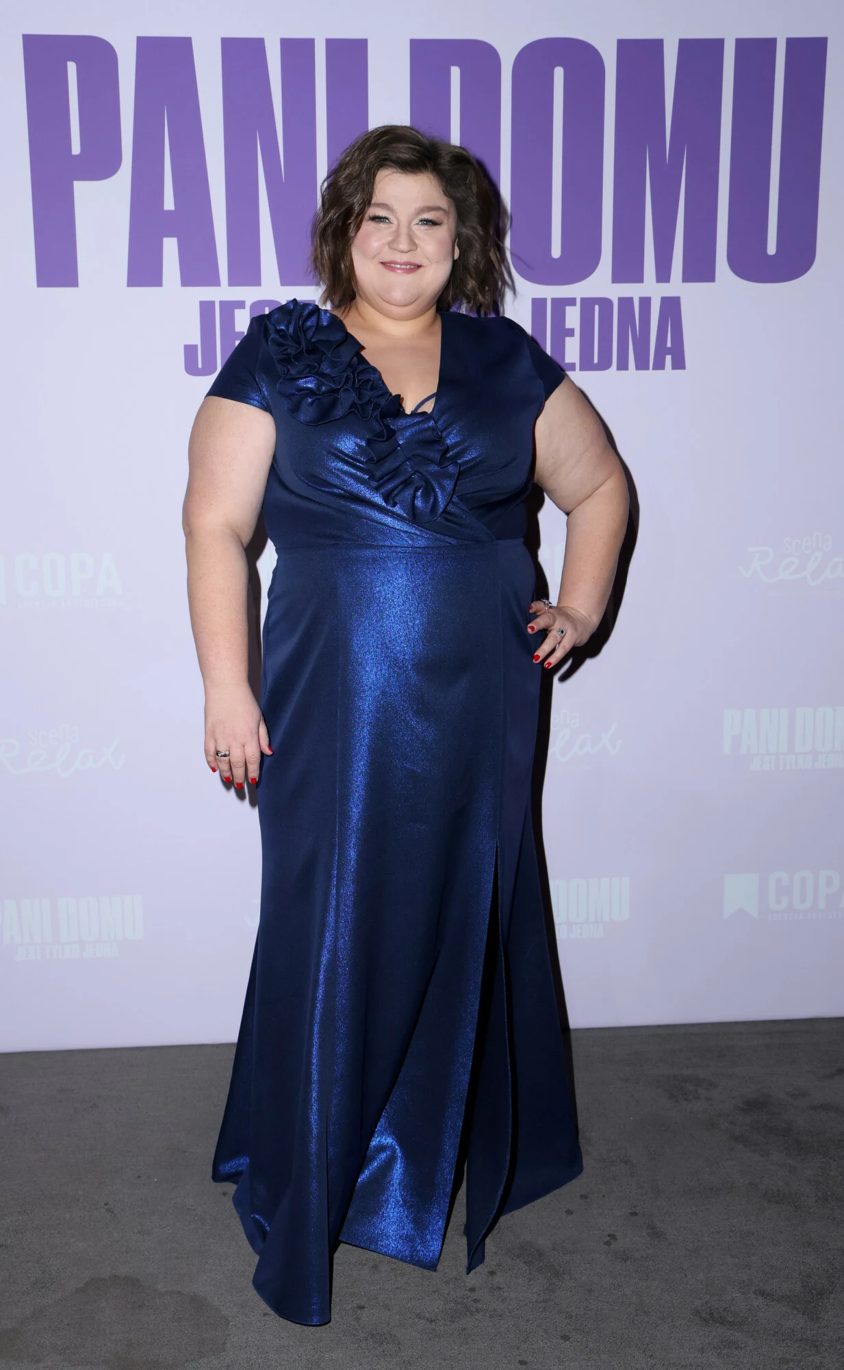 Kobieta w ciemnoniebieskiej, połyskującej sukni z dekoracyjnymi kwiatami na ramieniu stoi na tle ścianki z dużym napisem w kolorze fioletowym.