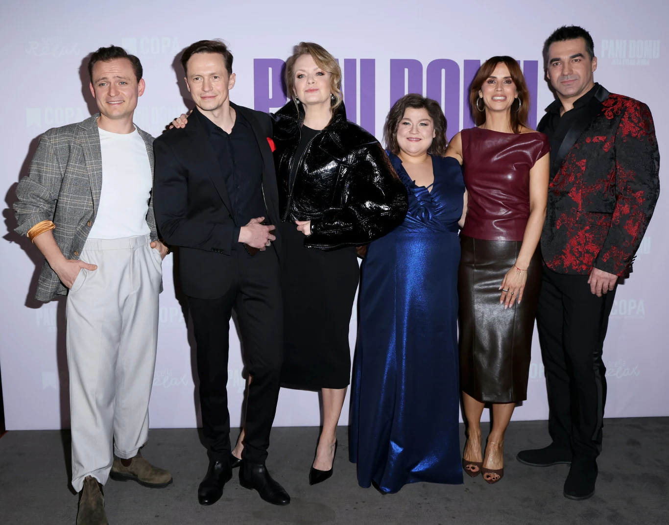 Grupa sześciu elegancko ubranych osób pozuje razem na tle fioletowej ścianki z logotypem, każda z nich prezentuje inną stylizację, od klasycznych garniturów po połyskujące sukienki i skórzaną kurtkę, wszyscy uśmiechnięci.