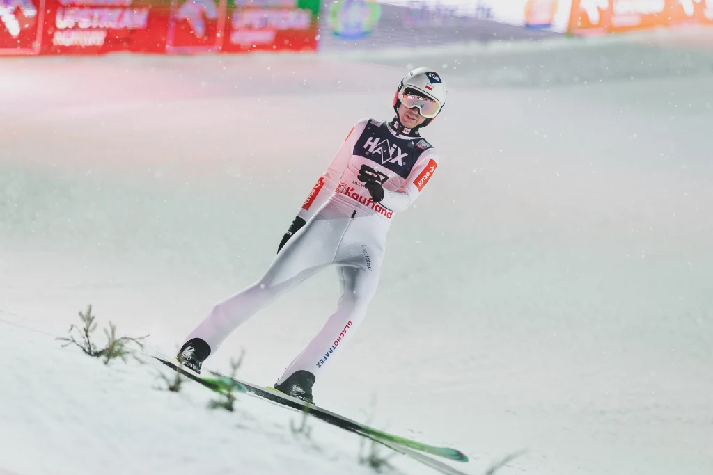 Kamil Stoch Skoczek narciarski w kombinezonie sportowym podczas lądowania na skoczni, dookoła rozpościera się zaśnieżona powierzchnia, w tle migoczą rozświetlone reklamy i tablice świetlne.