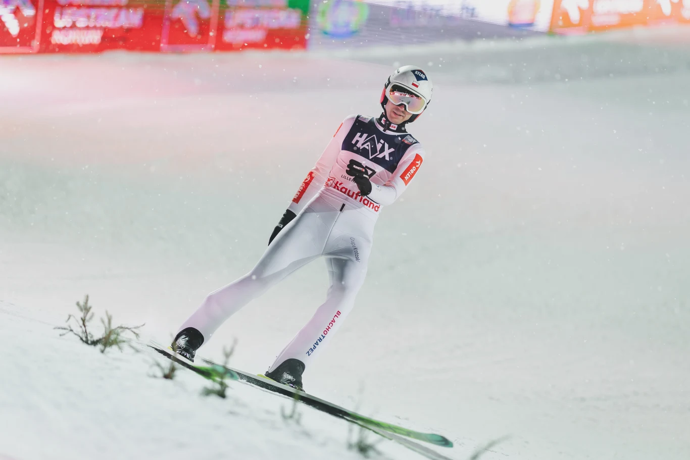 Kamil Stoch Skoczek narciarski w kombinezonie sportowym podczas lądowania na skoczni, dookoła rozpościera się zaśnieżona powierzchnia, w tle migoczą rozświetlone reklamy i tablice świetlne.