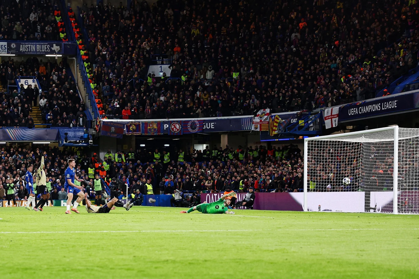 Piłka wpada do bramki podczas meczu piłkarskiego na stadionie, w tle tłum kibiców, bramkarz leżący na murawie po nieudanej interwencji, zawodnicy drużyn w dynamicznej akcji, atmosfera pełna emocji.