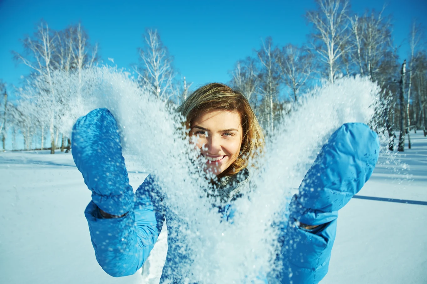 Los szykuje mocny zwrot. Te trzy znaki zodiaku mogą być naprawdę zaskoczone Uśmiechnięta kobieta w niebieskiej kurtce wyrzuca śnieg w powietrze na tle zimowego krajobrazu z ośnieżonymi drzewami i bezchmurnym niebem.