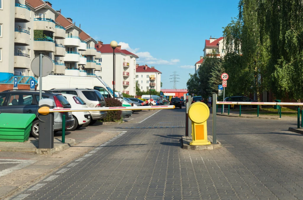 Szlaban na osiedlowej ulicy oddzielający parking od dalszej jazdy, po lewej stronie zaparkowane samochody oraz bloki mieszkalne z balkonami, po prawej stronie zieleń i chodnik.