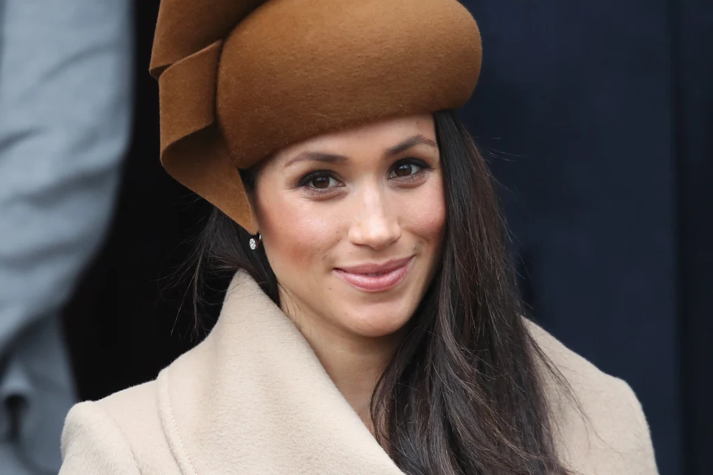 Meghan Markle skupiała na sobie uwagę podczas świątecznych spacerów z rodziną królewską Uśmiechnięta kobieta ubrana w elegancki płaszcz w kolorze beżowym oraz kapelusz w kolorze brązowym, z długimi ciemnymi włosami, znajdująca się na tle o ciemnej kolorystyce.