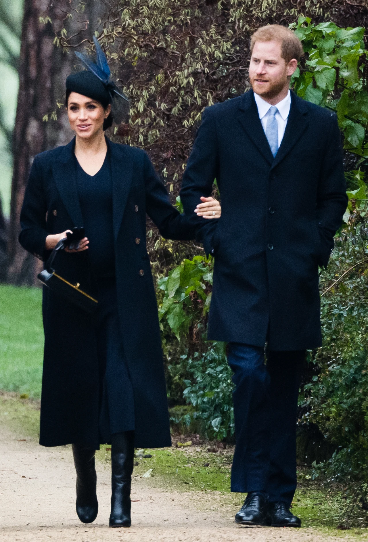 W 2018 roku Meghan Markle podczas świąt postawiła na ciemne barwy Elegancka para idzie razem alejką w otoczeniu zieleni. Kobieta ubrana w czarny płaszcz, sukienkę i ozdobny kapelusz, trzyma torebkę w jednej ręce, a drugą obejmuje mężczyznę. Mężczyzna ma na sobie ciemny płaszcz, garnitur oraz krawat, idzie pewnym krok...