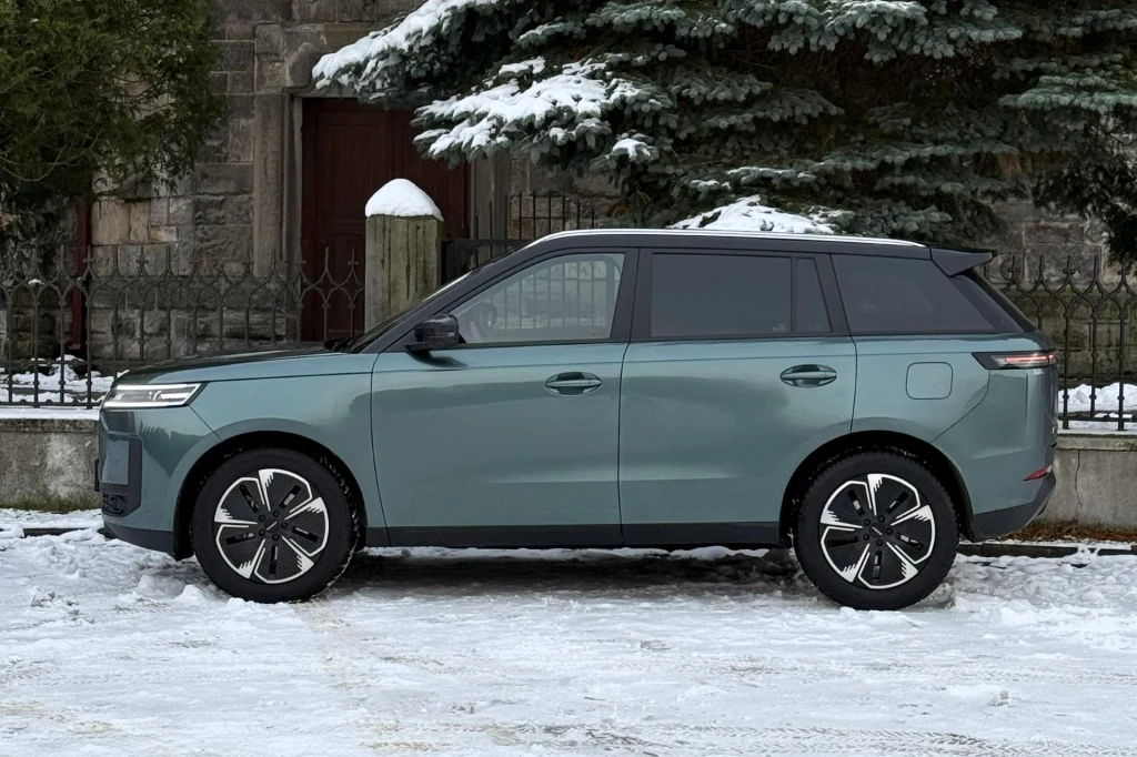 Zielony, nowoczesny SUV zaparkowany na zaśnieżonej drodze na tle kamiennego ogrodzenia i świerków pokrytych śniegiem.