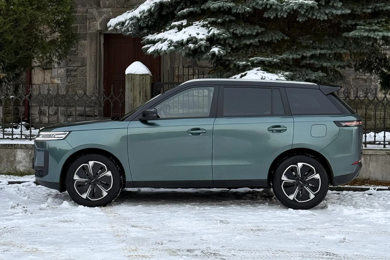 Zielony, nowoczesny SUV zaparkowany na zaśnieżonej drodze na tle kamiennego ogrodzenia i świerków pokrytych śniegiem.