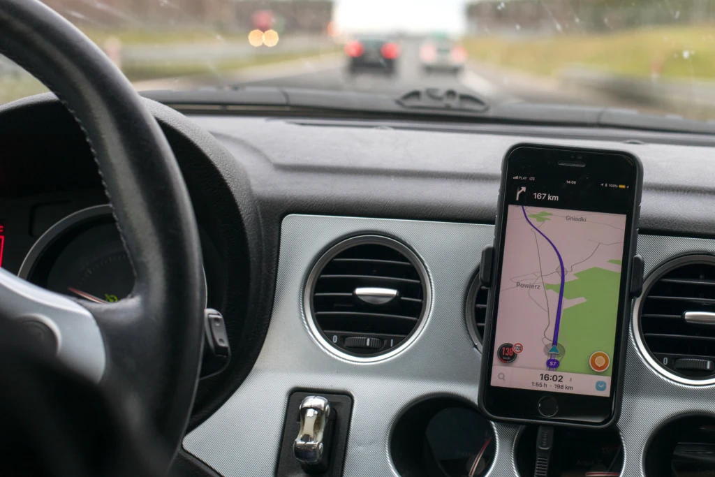 Środek samochodu z widoczną kierownicą i deską rozdzielczą, na której zamontowany jest smartfon z aplikacją nawigacyjną włączoną i wyznaczającą trasę. W tle widoczna rozmyta droga i samochody.