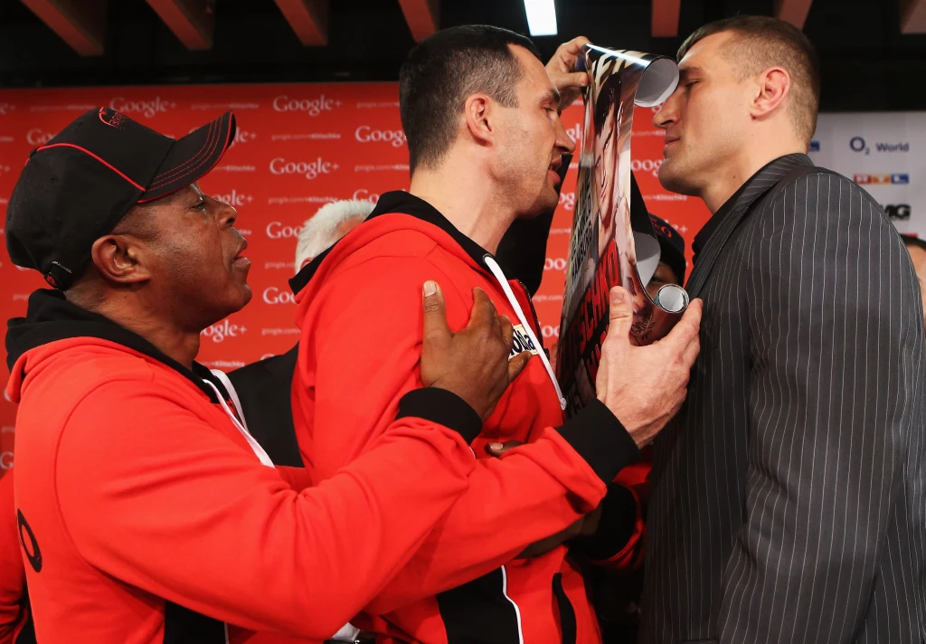 Władimir Kliczko i Mariusz Wach, 05.11.2012 r. Dwóch mężczyzn stoi twarzą w twarz podczas oficjalnego spotkania sportowego, jeden z nich trzyma plakat przy twarzy przeciwnika, obok stoi trener w czerwonej kurtce, a tło wypełniają czerwone i białe bannery reklamowe.