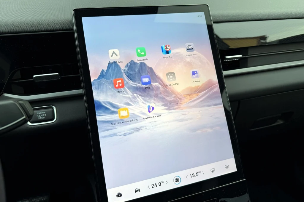 Duży dotykowy ekran multimedialny w samochodzie z interfejsem prezentującym aplikacje i ustawienia klimatyzacji na tle widoku górskiego.