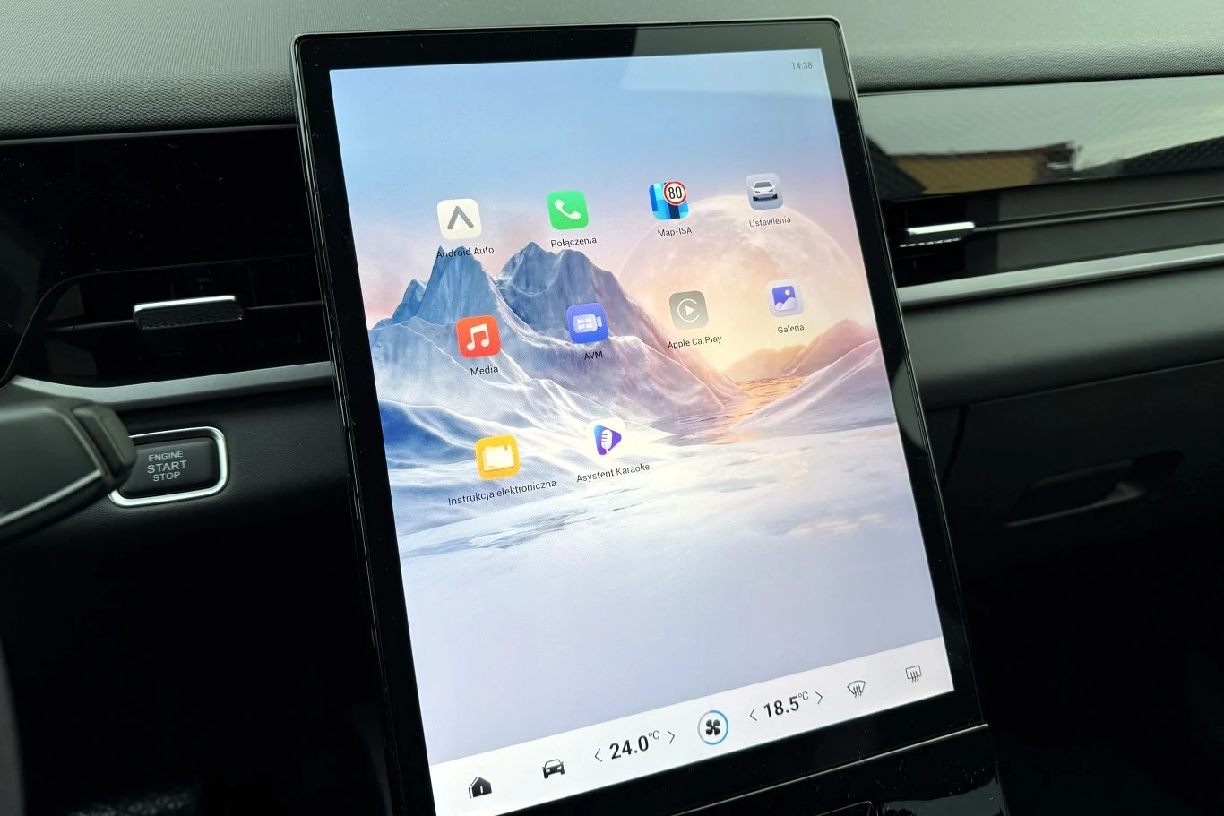 Duży dotykowy ekran multimedialny w samochodzie z interfejsem prezentującym aplikacje i ustawienia klimatyzacji na tle widoku górskiego.