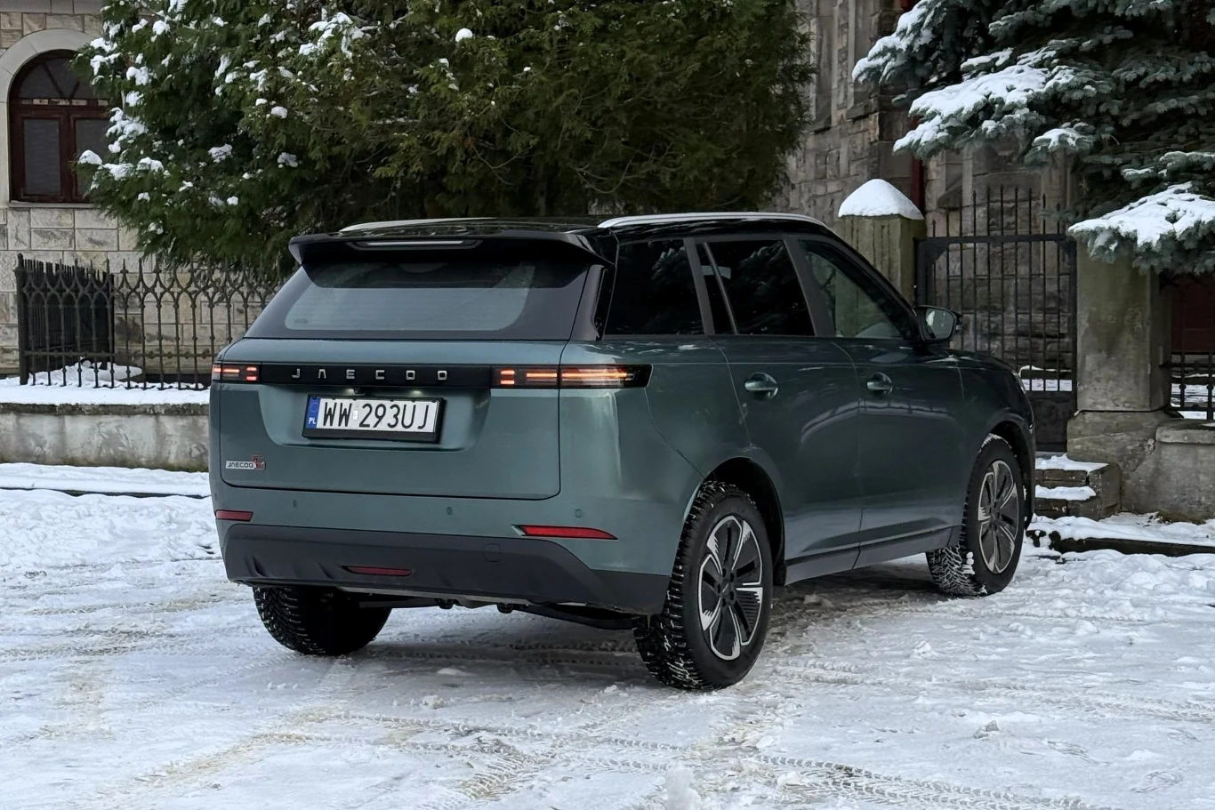 Zielony SUV marki Jaecoo zaparkowany na zaśnieżonym podjeździe, otoczony drzewami i murami budynku, z widoczną rejestracją oraz charakterystycznym, nowoczesnym designem tylnej części pojazdu.
