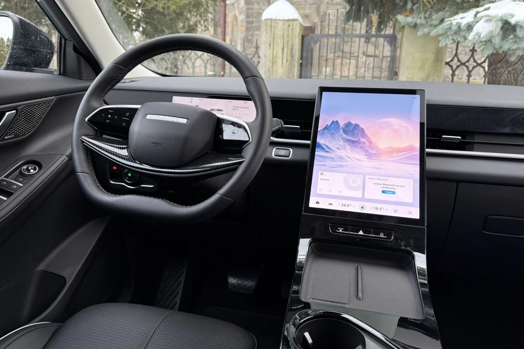 Nowoczesne wnętrze samochodu z dużym cyfrowym panelem dotykowym na środkowej konsoli, cyfrowymi zegarami za kierownicą oraz czarną tapicerką. Na ekranach widoczne są interfejsy multimedialne.