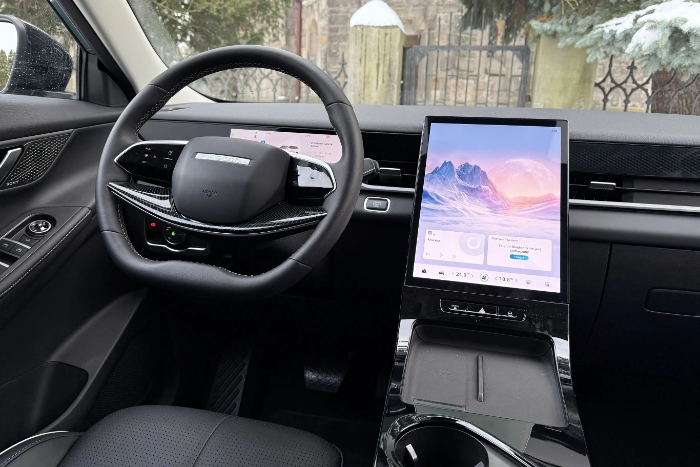 Nowoczesne wnętrze samochodu z dużym cyfrowym panelem dotykowym na środkowej konsoli, cyfrowymi zegarami za kierownicą oraz czarną tapicerką. Na ekranach widoczne są interfejsy multimedialne.