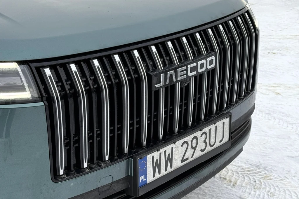Przód samochodu z dużym, pionowym chromowanym grillem marki Jaecoo oraz widoczną polską tablicą rejestracyjną na tle śnieżnego podłoża.