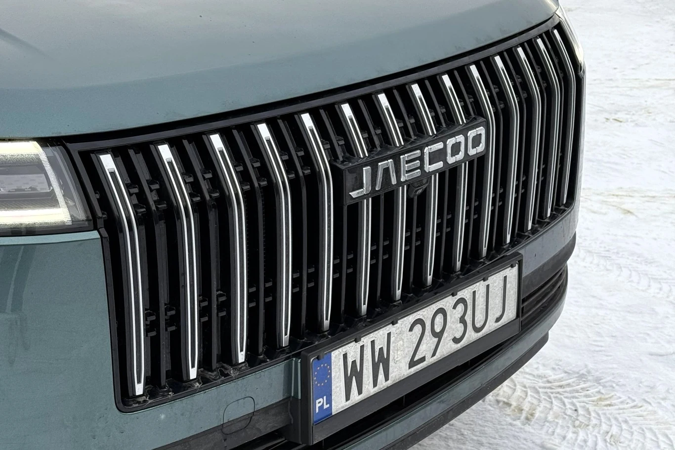 Przód samochodu z dużym, pionowym chromowanym grillem marki Jaecoo oraz widoczną polską tablicą rejestracyjną na tle śnieżnego podłoża.