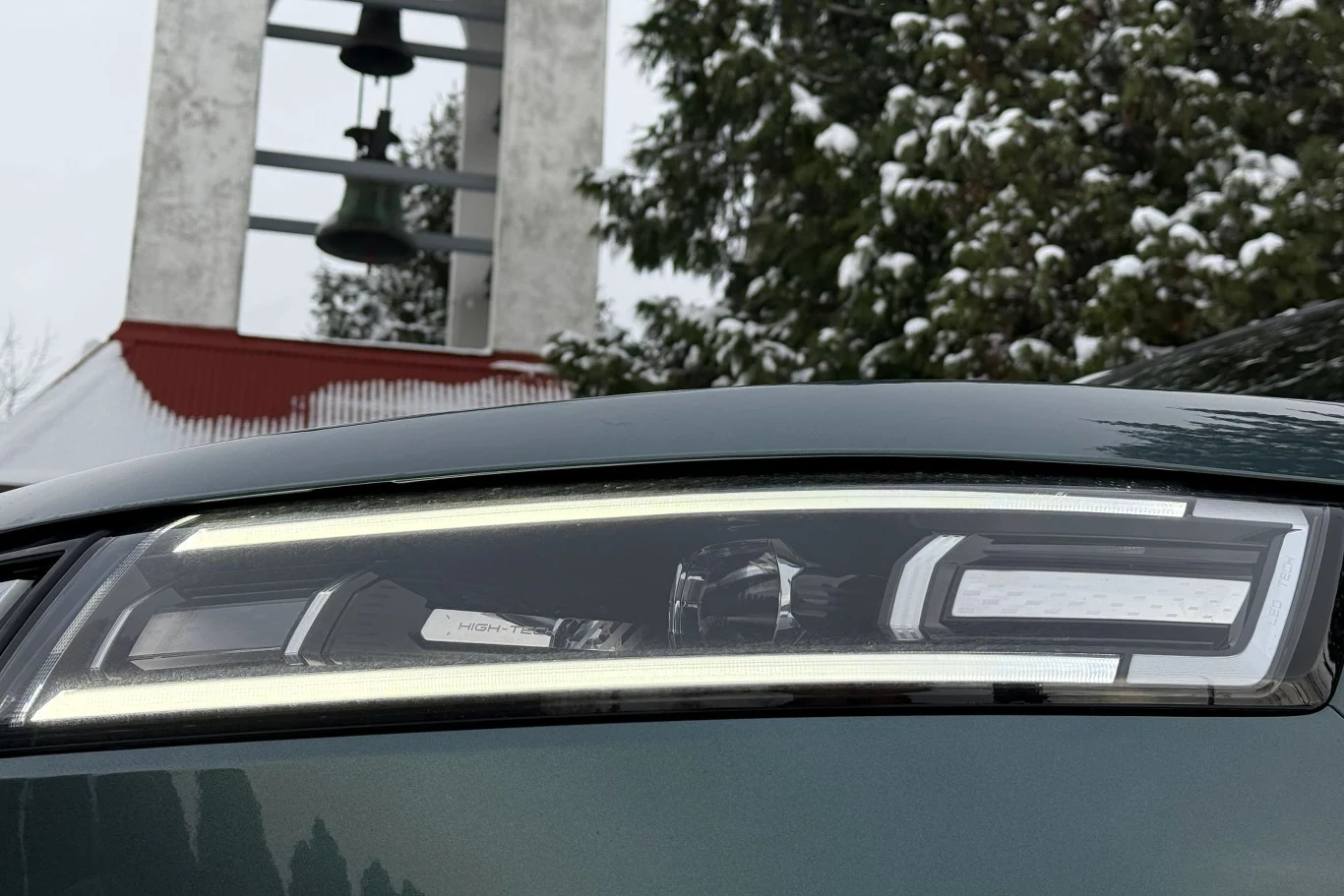 Nowoczesny reflektor samochodowy na tle zaśnieżonych drzew oraz dzwonnicy z dzwonami.