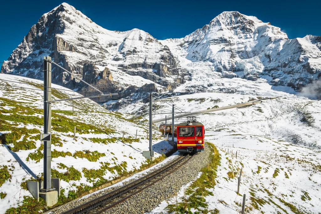 Słynny szwajcarski Glacier Express zabiera pasażerów w podróż po dzikich i zaśnieżonych zakątkach Alp Czerwony pociąg jadący po wąskiej torach górskich otoczonych ośnieżonymi szczytami alpejskimi przy bezchmurnym, niebieskim niebie.