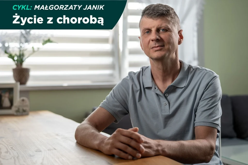 Siedzący przy stole mężczyzna w szarej koszulce polo, patrzący spokojnie przed siebie w jasnym, domowym wnętrzu. W tle roślina doniczkowa na parapecie i częściowo zasłonięte żaluzje.