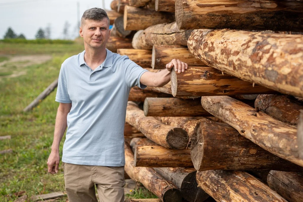 Mężczyzna w jasnoniebieskiej koszulce polo stoi przy dużym stosie ułożonych drewnianych bali, w tle widoczna zielona trawa i odległe drzewa.