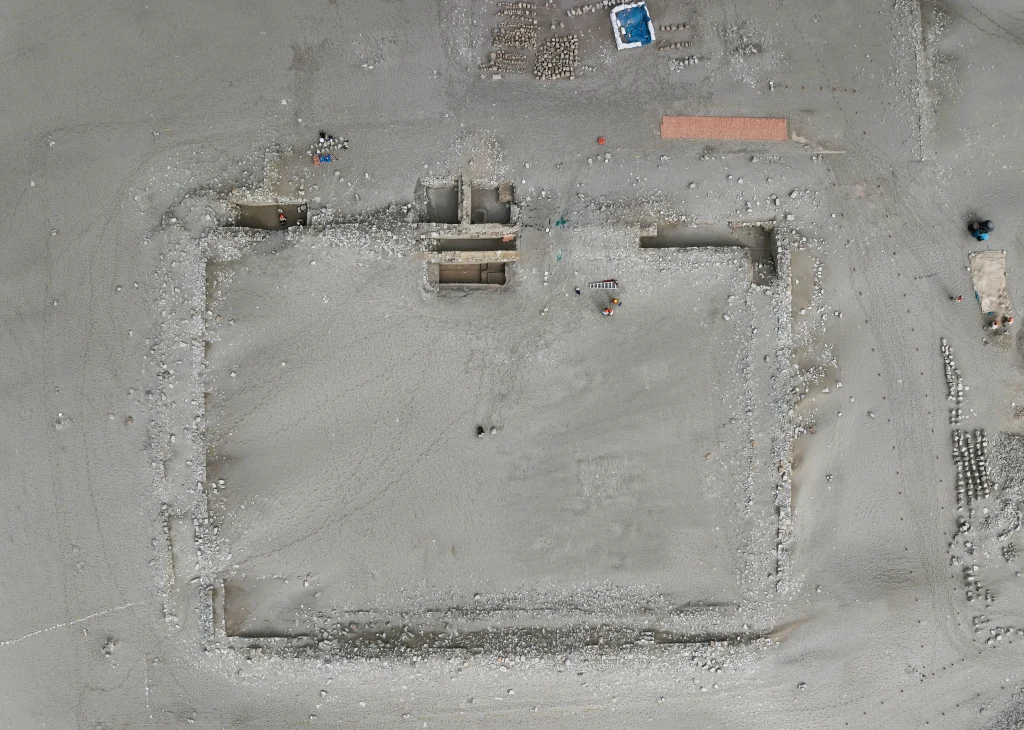 Ruiny prostokątnej budowli ukazane z lotu ptaka, otoczone szarym terenem z drobnymi elementami archeologicznymi wokół. Wewnątrz widoczne są pozostałości murów i zagłębień, a na zewnątrz drobne przedmioty i stanowiska pracy.