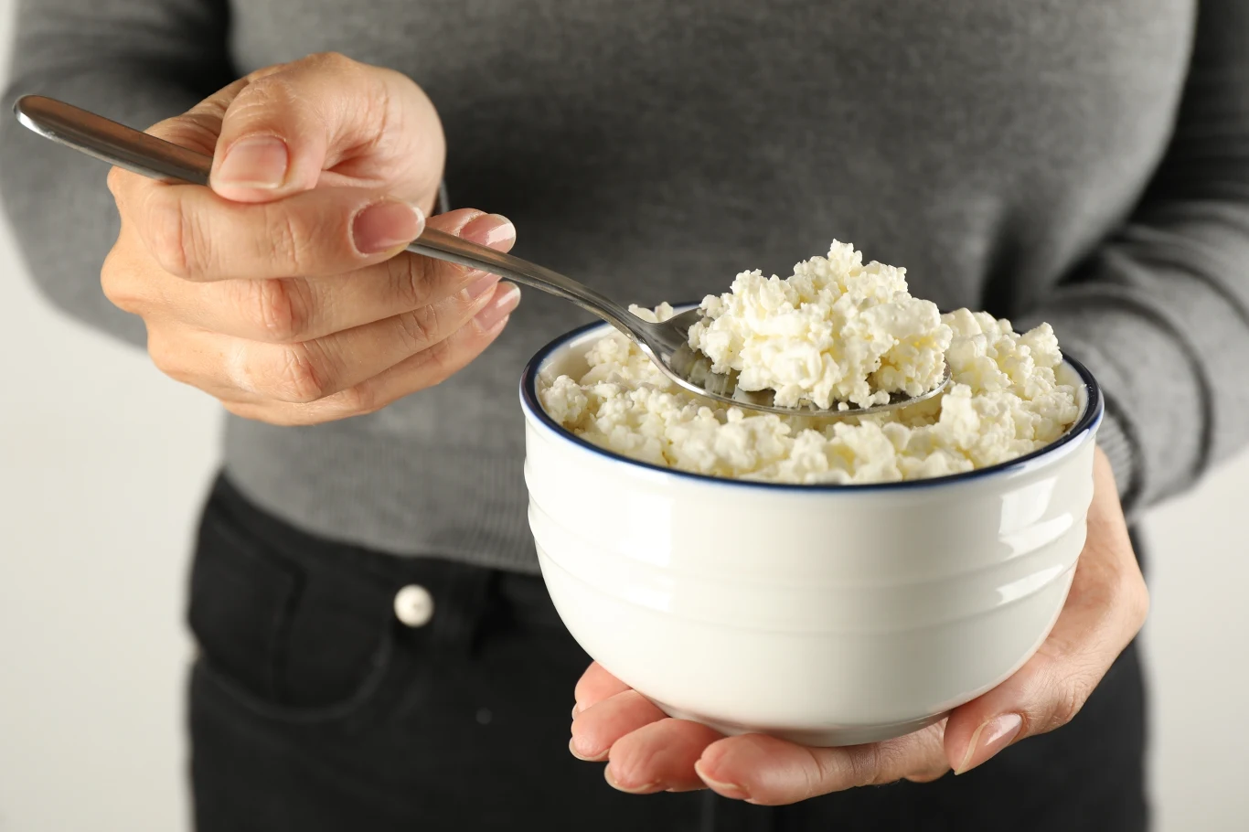 Osoba w szarej bluzce trzyma białą miskę z twarogiem, nabierając go łyżką.