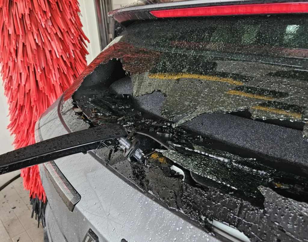 Siła była tak duża, że mechanizm wycieraczki został wyrwany z mocowania. Naprawa polega na wymianie całej tylnej klapy Rozbita tylna szyba samochodu, pośród której widać szczątki szkła na powierzchni klapy oraz tylnej wycieraczce. W tle znajduje się fragment czerwonych szczotek automatycznej myjni samochodowej.
