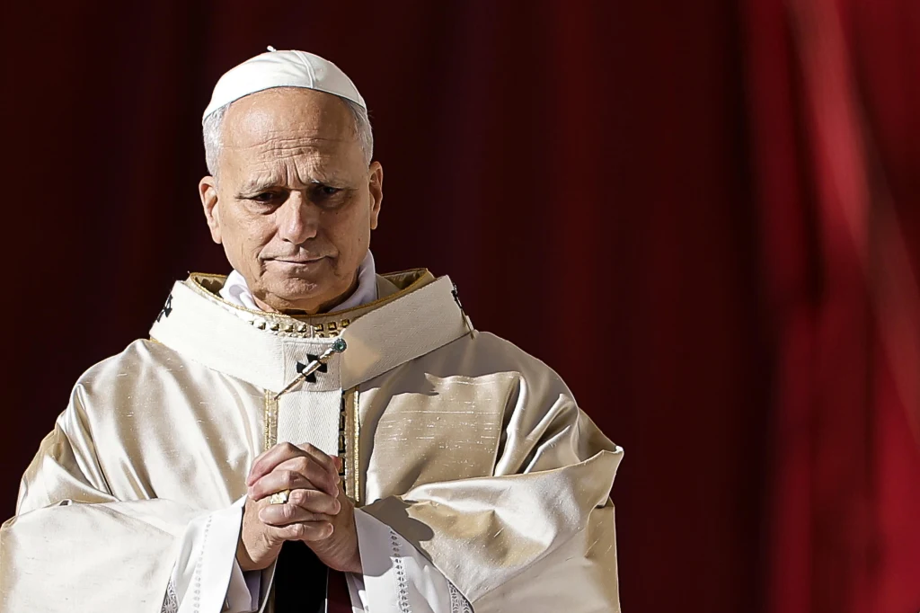 Mężczyzna w liturgicznych szatach katolickich z białą piuską modli się ze złożonymi dłońmi na tle czerwonej zasłony.