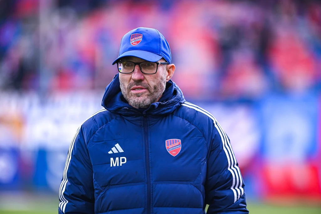 Marek Papszun Mężczyzna w okularach i czapce z daszkiem ubrany w niebieską kurtkę sportową z logo klubu oraz inicjałami MP, stoi na stadionie z rozmytym tłem w barwach niebiesko-czerwonych.